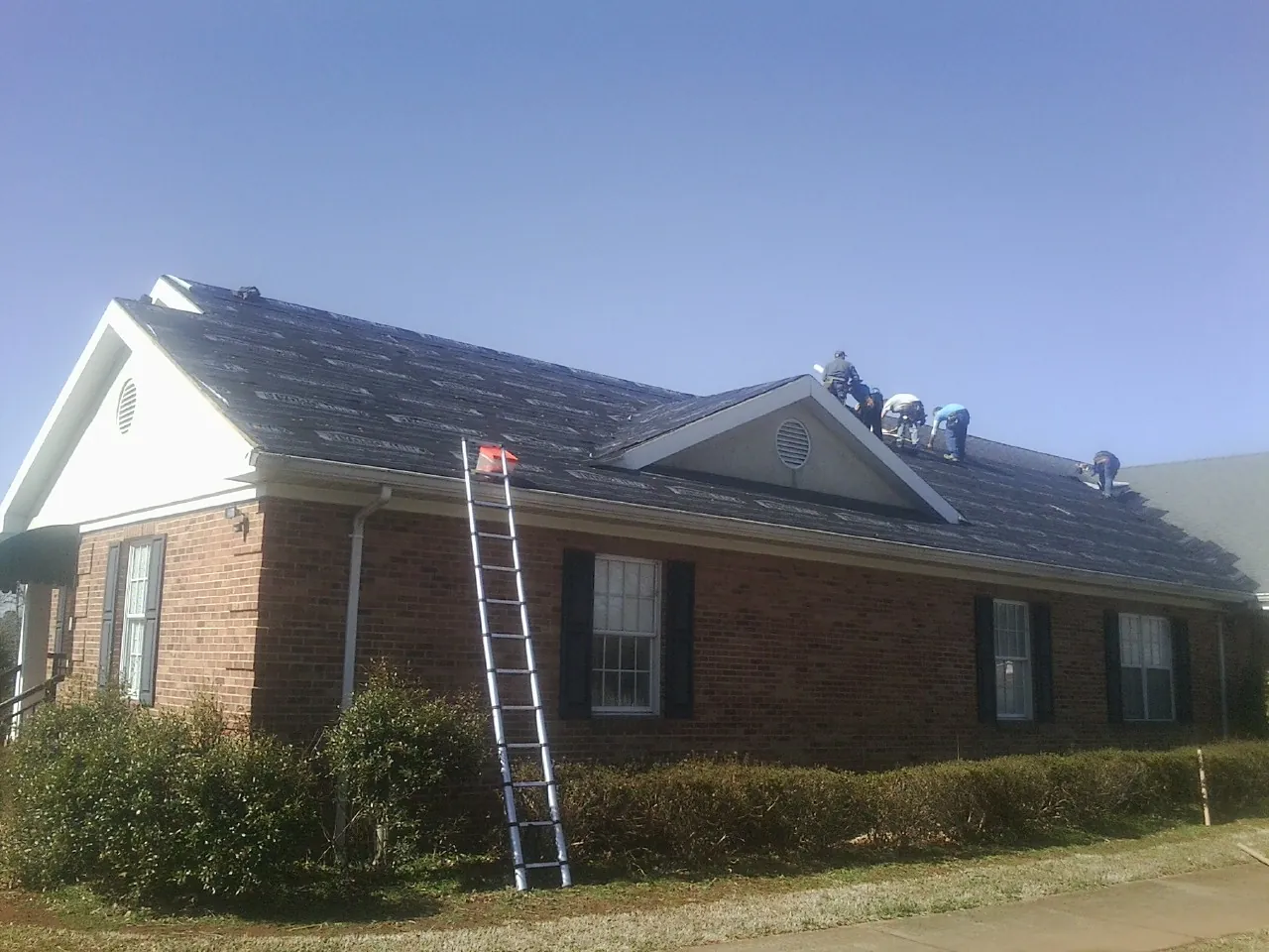 Crew tearing shingles off a brick ranch roof with a ladder set — Metal Roof Repair work by our crew in Carman