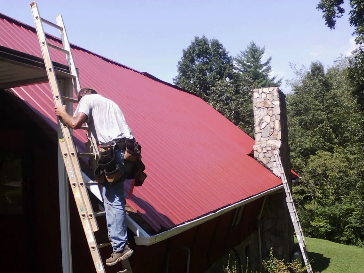 Roofer on a ladder beside a red metal roof and stone chimney — Soffit & Fascia Repair by Roofing Brothers in Carman
